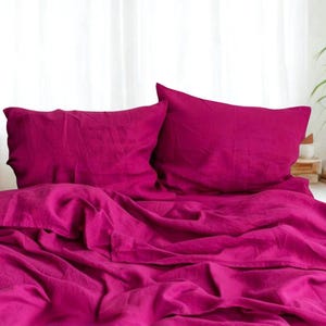 May include: A bed set featuring a vibrant fuchsia color. The set includes two pillowcases and a folded sheet, all made of a soft, textured fabric. The bedding is arranged on a bed, creating a cozy and inviting atmosphere.