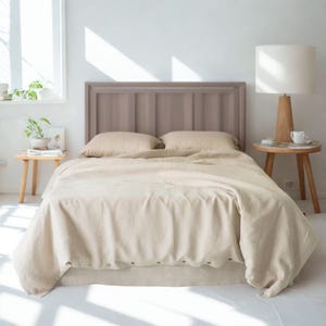 May include: A neutral-toned bedroom set featuring a bed with a wooden headboard, pillows, and a duvet cover. The bedding is a light beige color, creating a calming atmosphere. A bedside table with a lamp and a small table with plants are also visible.