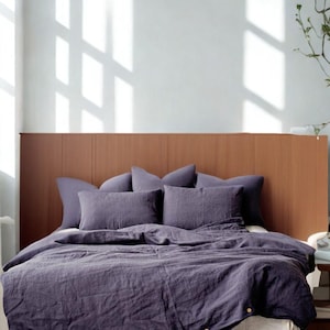 Op de afbeelding: Een bed met een houten hoofdbord en donkerpaars beddengoed. Het bed heeft een witte basis en meerdere kussens. Het beddengoed omvat een dekbedovertrek en kussenslopen in bijpassende kleur. De kamer heeft natuurlijk licht.