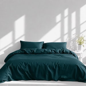 May include: A deep teal bedding set featuring a solid-colored duvet cover and two matching rectangular pillowcases. The fabric has a smooth, matte finish and is draped neatly over a bed frame, creating a clean and uniform appearance.