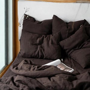 May include: A bed with dark brown linen bedding, including pillows and a duvet. A white headboard is visible, and a magazine rests on the bed. The scene is illuminated by natural light from a nearby window, creating a cozy atmosphere.