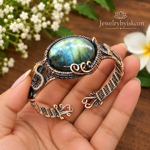May include: A wire-wrapped cuff bracelet featuring a large, oval-shaped labradorite gemstone with blue and green iridescence. The bracelet is crafted from dark oxidized copper wire, showcasing intricate woven patterns and decorative scrollwork ends.