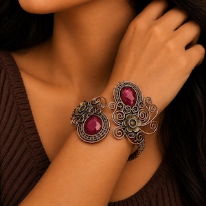 May include: A copper-toned wire-wrapped cuff bracelet featuring two faceted deep magenta oval gemstones. The metalwork includes intricate swirling wire patterns and small decorative flower-shaped metal accents integrated into the design.