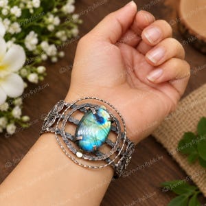 May include: A wide cuff bracelet featuring a large, oval-shaped labradorite gemstone with blue and green iridescence. The metal setting is oxidized silver with a textured, braided wire design and a circular frame surrounding the central stone.