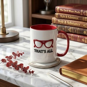 May include: White ceramic mug with a red interior and handle, featuring a red glasses design and the text "THAT'S ALL." The mug sits on a round, light-colored coaster. Books and a pen are in the background.