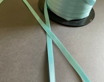 May include: Two spools of light blue satin ribbon. One spool is unwound and the ribbon is laying flat on a gray surface.