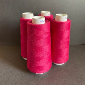 Könnte beinhalten: Vier Spulen leuchtend pinkfarbenes Garn. Jede Spule hat einen weißen Boden und Deckel, und das Garn ist eng gewickelt. Die Spulen sind eng beieinander angeordnet und zeigen die satte Farbe und Textur des Garns.