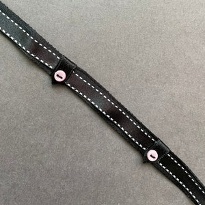 Op de afbeelding: Zwart lint met witte stiksels en twee roze knopen. Het lint ligt op een grijze achtergrond.