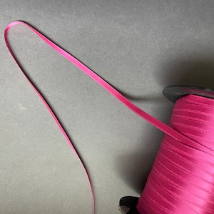 May include: A spool of pink satin ribbon with a single strand of ribbon extending from the spool.