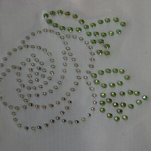 May include: A white canvas with a design of sparkling rhinestones. The design features a spiral and leaf-like shapes, with green rhinestones forming the leaves and clear rhinestones for the spiral. The canvas has a grid-like texture.