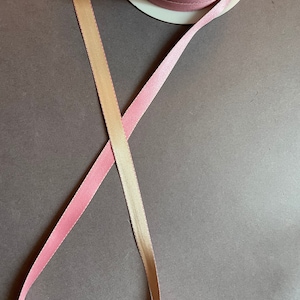 May include: Two spools of ribbon, one pink and one cream, are crossed over each other on a dark grey surface. The ribbon is satin and has a subtle sheen.