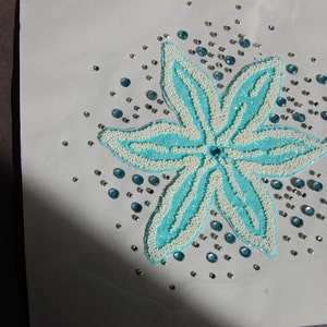 May include: A floral design with a turquoise and white beaded pattern. The flower has six petals and a central blue gem. Scattered around the flower are small silver and blue gems, creating a sparkling effect. The design is on a white background.