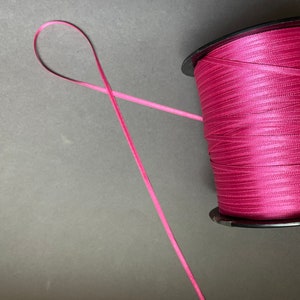 May include: A spool of pink satin ribbon with a length of ribbon extending from the spool.
