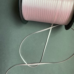 May include: A spool of pink satin ribbon with thin white stripes. The ribbon is wound around a black spool and a length of ribbon is unraveled.