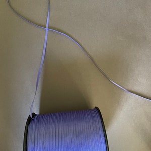 May include: A spool of lavender satin ribbon with a black plastic spool. The ribbon is wound around the spool and a length of ribbon is hanging off the spool.