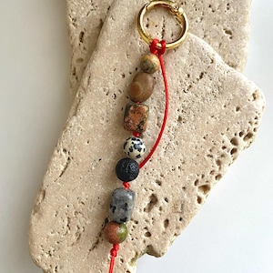 May include: A keychain featuring a gold-colored clasp and a strand of colorful, natural stones. The stones include brown, speckled, and black varieties, strung on a vibrant red cord. The keychain is displayed against a textured, light-colored stone background.