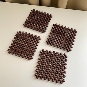 Op de afbeelding: Een set van vier vierkante onderzetters gemaakt van donkerbruine houten kralen in een rasterpatroon. Elke onderzetter heeft een flexibel, onderling verbonden ontwerp dat bestaat uit kleine, gepolijste, bolvormige houten kralen.