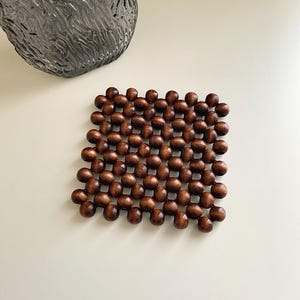 May include: A square trivet constructed from dark brown wooden beads arranged in a grid pattern. The beads are connected to form a flexible, open-work mat with a smooth, polished finish, showcasing the natural grain of the wood in each bead.