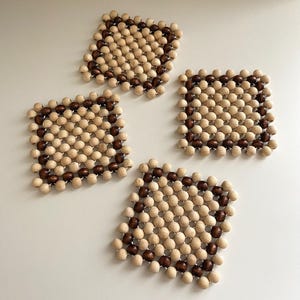 May include: A set of four square coasters constructed from small wooden beads strung together. Each coaster features a grid pattern with a border of dark brown beads surrounding a centre of light beige beads, creating a distinct contrast.