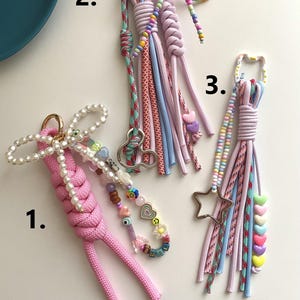 Può includere: Tre portachiavi fatti a mano con cordoncini colorati e dettagli di perline. Uno è rosa con un fiocco di perle e un ciondolo di perline. Un altro ha un ciondolo a forma di stella e cordoncini pastello.
