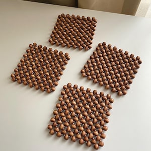 May include: A set of four square-shaped coasters constructed from numerous small, round, brown wooden beads linked together in a grid pattern. Each coaster features a uniform texture created by the interconnected bead arrangement in a flat design.