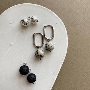 Może przedstawiać: Kolekcja trzech par kolczyków prezentowanych na białej, zakrzywionej powierzchni. Kolczyki posiadają srebrny metal i różne kamienne koraliki, w tym teksturowane srebro, nakrapiane biało-czarne i czarny kamień lawowy.