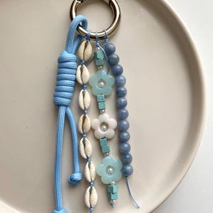 Pode incluir: Um porta-chaves azul claro com um fecho prateado. O porta-chaves apresenta uma corda azul, conchas brancas, contas turquesas, contas de flores brancas e contas de pedras preciosas azuis. O porta-chaves está sobre um prato branco.