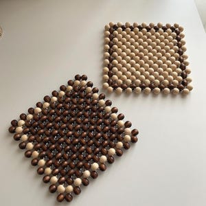 Può includere: Due sottobicchieri di perline di legno. Uno è quadrato, l'altro a forma di diamante. Entrambi sono fatti di piccole perline rotonde, marrone chiaro e scuro. I sottobicchieri sono progettati per proteggere le superfici da calore e umidità.