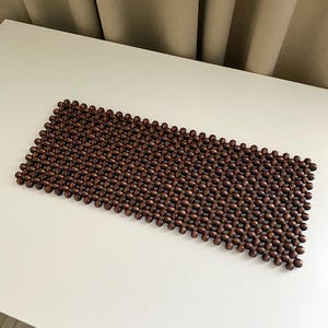 May include: A rectangular brown beaded mat made of small, dark wooden beads. The beads are strung together to create a textured surface. The mat is placed on a white surface.