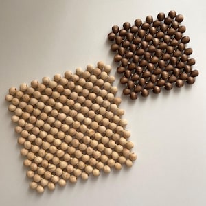 Può includere: Due sottopentola quadrati con perline di legno. Uno è di colore chiaro, l'altro è marrone scuro. Le perline sono disposte vicine, creando una superficie strutturata. Questi sottopentola possono essere usati come sottopentola o elementi decorativi.