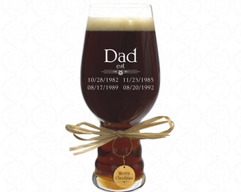 May include: A clear glass beer stein with a brown liquid inside. The stein is engraved with the text "Dad est. 10/28/1982 11/23/1985 08/17/1989 08/20/1992". A gold tag with the text "Merry Christmas" is attached to the stein.