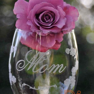 May include: A clear glass wine glass with the word "Mom" etched on the side. The glass is decorated with small, etched flowers. A single purple rose is placed inside the glass.