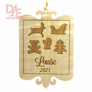 May include: Wooden Christmas ornament with engraved reindeer, sleigh, teddy bear, mitten, and Christmas tree designs. The name "Louise" and the year "2021" are also engraved on the ornament. The ornament has a decorative border and a ribbon for hanging.