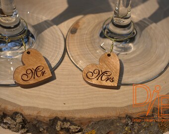 May include: Two wooden heart-shaped wine glass charms with the words "Mr." and "Mrs." engraved on them. The charms are attached to gold-colored metal rings.