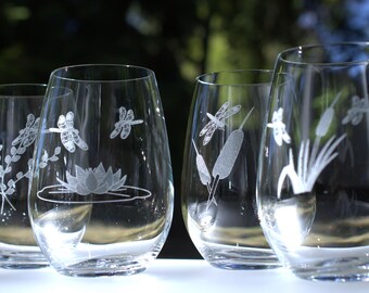 May include: Four clear glass wine glasses with etched designs of dragonflies, cattails, and a water lily. The glasses are on a white surface.