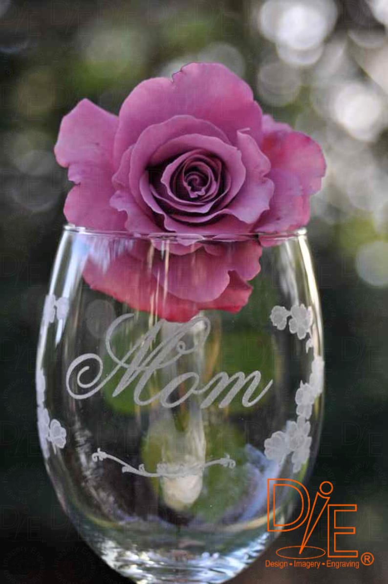May include: A clear glass wine glass with the word "Mom" etched on the side. The glass is decorated with small, etched flowers. A single purple rose is placed inside the glass.