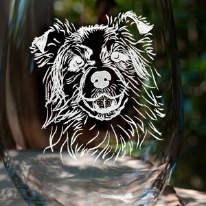 K&ouml;nnte beinhalten: Ein klares Weinglas aus Glas mit einem wei&szlig;en, ge&auml;tzten Design eines Hundegesichts. Der Hund hat schwarzes Fell und schaut direkt auf den Betrachter.