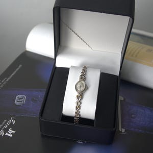 May include: A silver-toned analogue wristwatch with an oval face and a delicate link bracelet featuring heart-shaped accents. The watch is displayed on a white cushion inside a black hinged presentation box with a white interior lining.