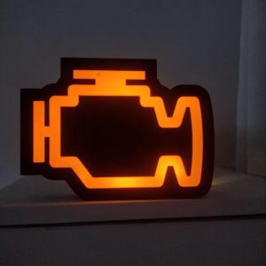 Peut inclure: Une enseigne noire et plate en forme de moteur avec une lumière orange brillante qui illumine le contour interne du symbole du moteur. Le design imite l'apparence d'un voyant de contrôle moteur de tableau de bord dans un environnement sombre.