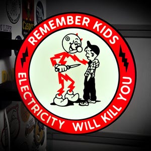 May include: A circular, illuminated wall sign with a red border and white center. The text "REMEMBER KIDS ELECTRICITY WILL KILL YOU" is printed in black. A graphic shows a red lightning bolt character holding a knife toward a young boy.