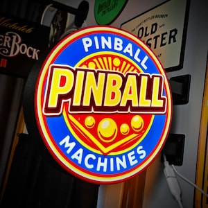 May include: A circular, illuminated wall sign featuring a blue border with the white text PINBALL MACHINES. The center has a red background with yellow spheres and the large yellow text PINBALL with a dark red outline and a sunburst graphic.