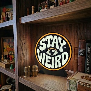 May include: A circular illuminated sign with a black frame and a glowing white centre featuring the text STAY WEIRD in a stylised font, with an eye graphic positioned between the two words, set on a black triangular base on a wooden shelf.
