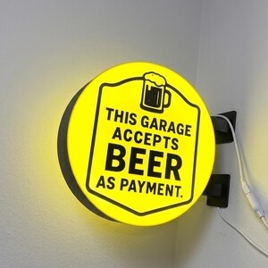 May include: A circular, illuminated wall-mounted sign with a bright yellow face and black border. It features a graphic of a beer mug and the black text: THIS GARAGE ACCEPTS BEER AS PAYMENT. The sign is attached to a black bracket with a white power cord.