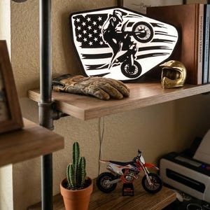 May include: A backlit sign featuring a black silhouette of a motocross rider performing a wheelie against a white American flag background. The sign sits on a wooden shelf next to a pair of worn leather gloves and a small gold-colored helmet model.