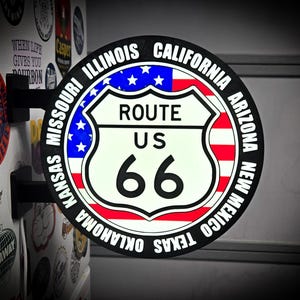 May include: A circular, double-sided wall-mounted sign featuring the Route 66 shield logo over a red, white, and blue American flag background. The black border lists the states of Illinois, Missouri, Kansas, Oklahoma, Texas, New Mexico, Arizona, and California.