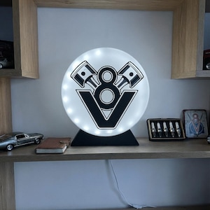 Può includere: Una lampada LED circolare con una grafica nera di un motore V8 che mostra due pistoni e il numero otto. La luce è montata su una base triangolare nera ed emette un tenue bagliore bianco da dietro il pannello circolare traslucido.