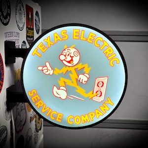 May include: A circular, illuminated wall-mounted sign featuring a light blue background with yellow text reading Texas Electric Service Company. A cartoon character with a lightning bolt body is depicted plugging into a white electrical outlet.