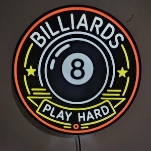 May include: A circular neon-style sign featuring a black 8-ball graphic in the center. The text BILLIARDS is arched across the top, and the text PLAY HARD is displayed on a banner at the bottom. The sign includes yellow stars and orange borders.