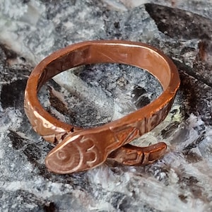 May include: A copper-coloured metal ring shaped like a coiled snake. The band features a textured surface with etched details, including a circular eye and segmented markings along the body, forming a wrap-around design that overlaps at the front.