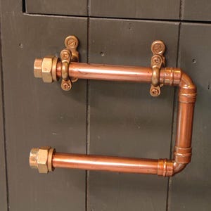 Könnte beinhalten: Zwei Handtuchhalter aus Kupferrohren, montiert auf einer dunkelgrünen Holzoberfläche. Jeder Halter verfügt über Messingbeschläge und eine rechtwinklige Biegung. Das Design hat eine industrielle, steampunk-Ästhetik.